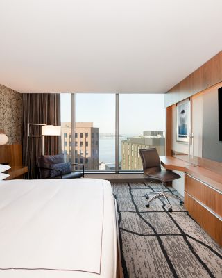 A modern hotel room with a large bed, a desk and chair, a flat-screen TV, and a view of buildings and a body of water through a window.