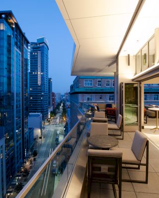 A modern city view from an outdoor terrace at dusk, with tables, chairs, and an open indoor area. Illuminated high-rise buildings line the street.
