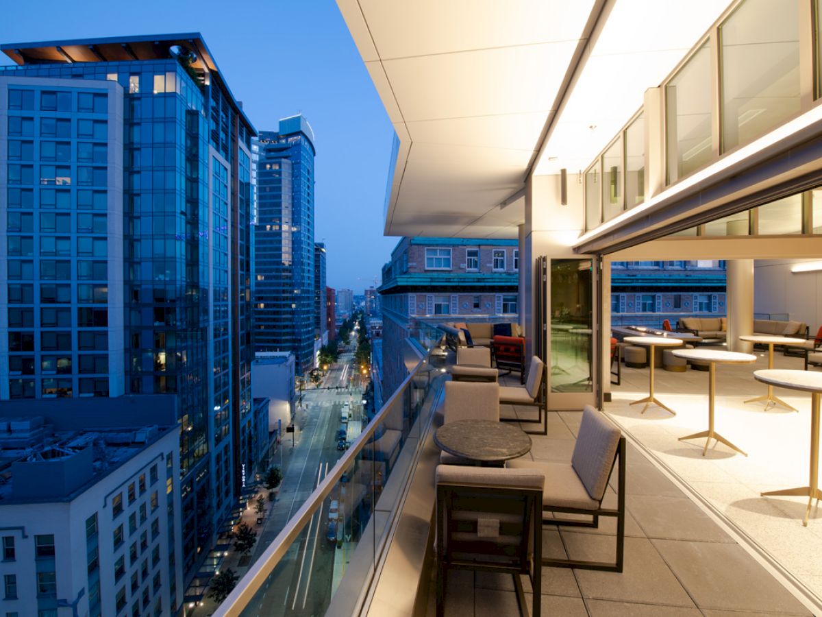A modern balcony with seating and tables, overlooking a cityscape with tall buildings, during the evening.