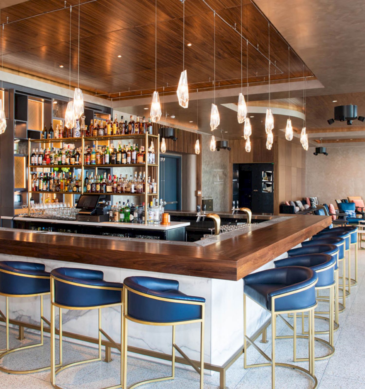 The image shows a modern bar with blue barstools, a well-stocked bar, and hanging lights. There is seating area in the background, creating a cozy ambiance.