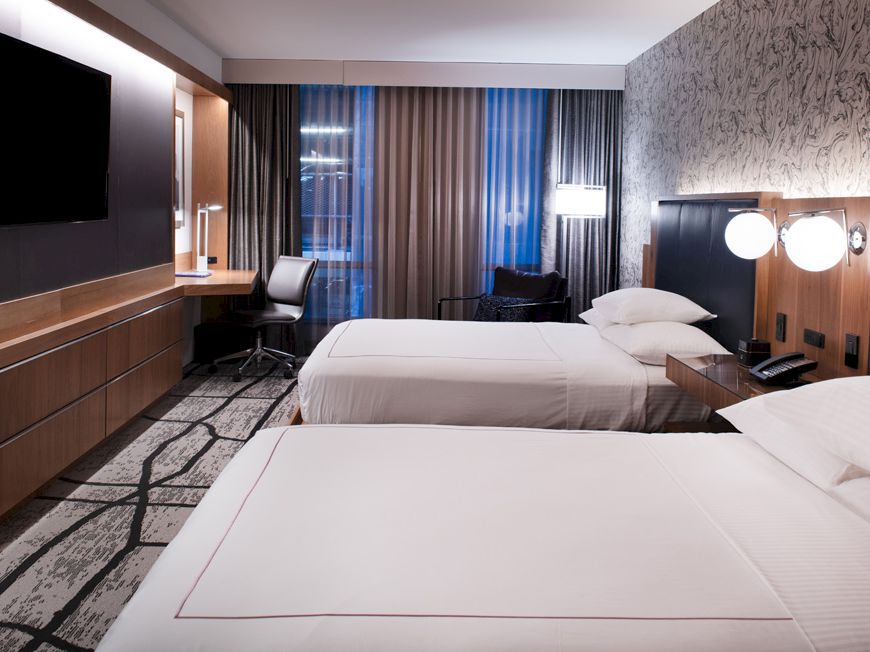 A modern hotel room with two neatly made beds, a large flat-screen TV, a desk with a chair, and a window with curtains.