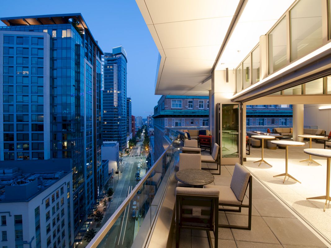 A modern balcony at dusk, featuring comfortable seating, small tables, and a cityscape view with tall buildings and a lit street below.
