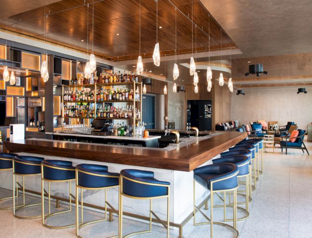 A modern bar with a wooden counter, blue bar stools, hanging lights, and a well-stocked liquor display behind it. Seating areas are in the background.