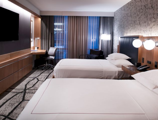 The image shows a modern hotel room with two neatly made beds, a flat-screen TV on the wall, a desk with a chair, and large windows with curtains.