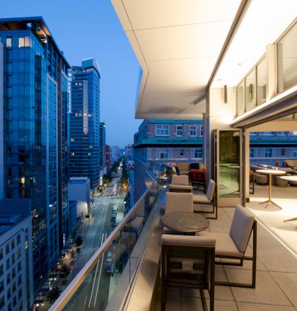 The image features a modern balcony overlooking a cityscape with tall buildings. The balcony has outdoor seating and tables.