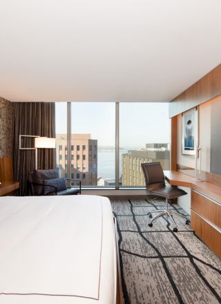 A modern hotel room with a large bed, wall-mounted TV, desk, chair, patterned rug, and floor-to-ceiling windows showing a cityscape view.