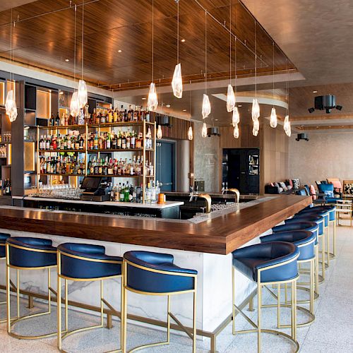 A modern bar with blue stools, a wooden countertop, overhead pendant lights, a well-stocked liquor shelf, and a lounge area in the background.