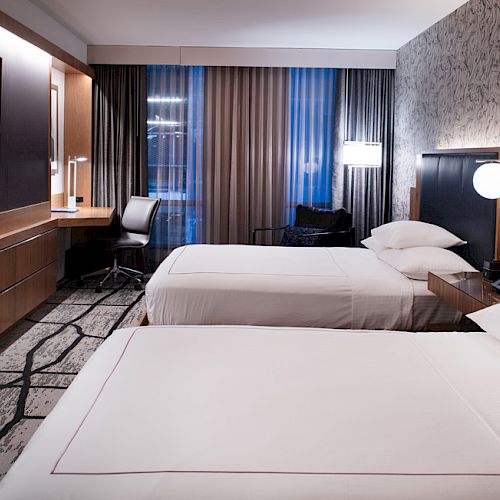 The image shows a modern hotel room with two neatly made beds, a desk with a chair, a flat-screen TV, and stylish decor.