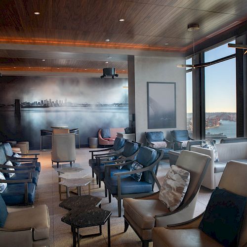 A modern lounge with comfortable seating, large windows, cityscape mural, and a view of a Ferris wheel and water outside, under a wooden ceiling.