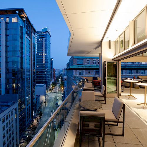 A modern balcony with seating overlooks a cityscape of tall buildings during dusk, featuring a glass railing and outdoor tables.