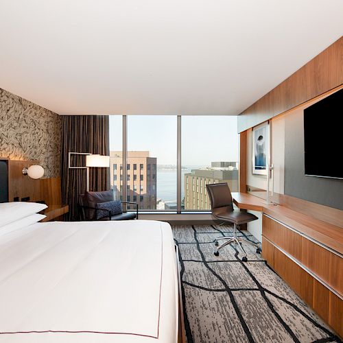 The image shows a modern hotel room with a large bed, desk, chair, flat-screen TV, and cityscape view through large windows.