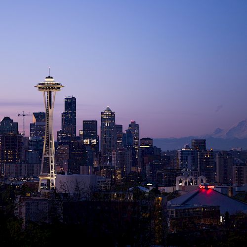 A view of the Seattle skyline at twilight, featuring the Space Needle and Mount Rainier in the background, with buildings illuminated against the evening sky.
