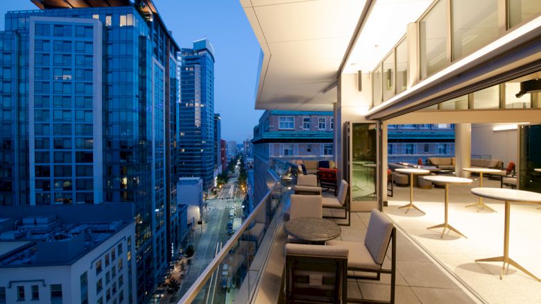 The image shows a modern balcony overlooking a city with tall buildings, featuring seating areas and tables, with the city lights visible in the evening.