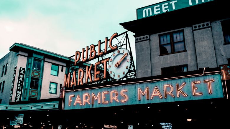 The image displays the entrance of a building labeled "PUBLIC MARKET" and "FARMERS MARKET" in neon lights, with a clock and additional signage.