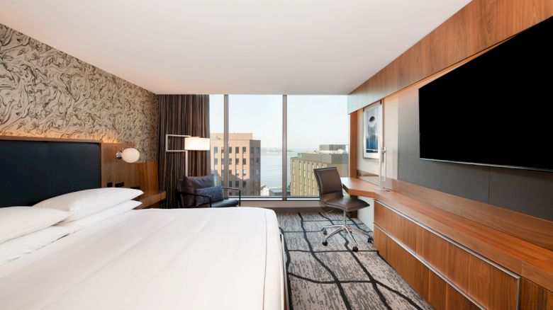 A modern hotel room with a large bed, sleek wooden furnishings, a desk with a chair, a large TV, and a window view of buildings and water.
