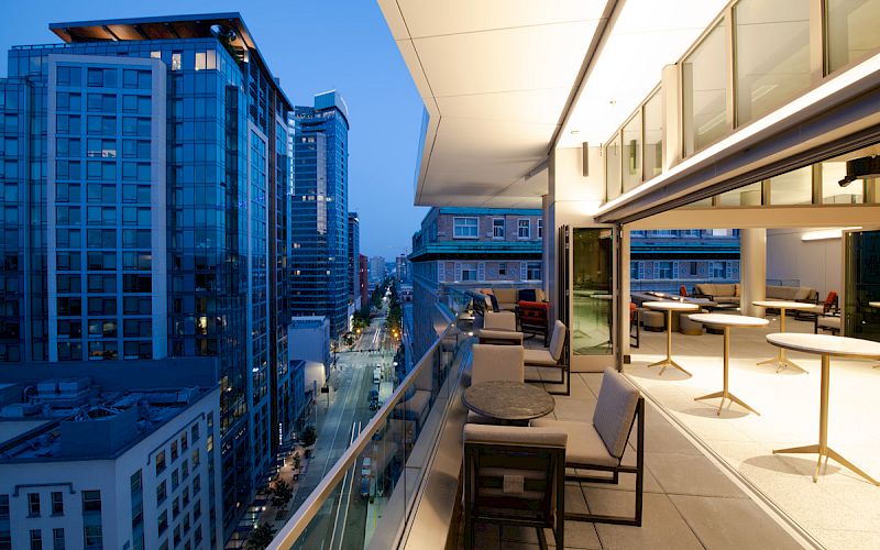 A modern balcony with seating overlooks a city street filled with tall buildings during twilight hours, creating an urban evening ambiance.