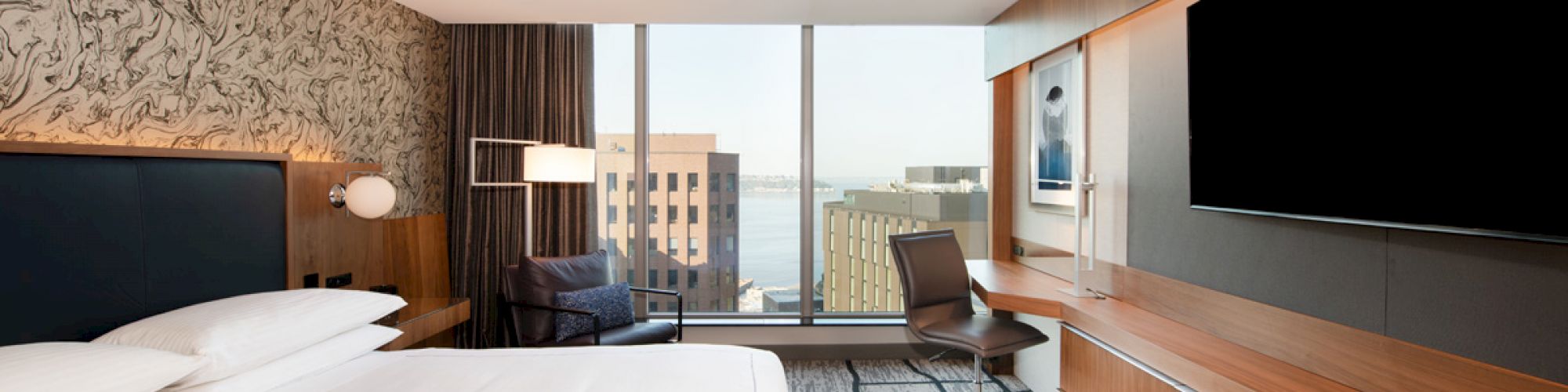 A modern hotel room with a large bed, a desk, a chair, a wall-mounted TV, and a view of buildings and water through the window.