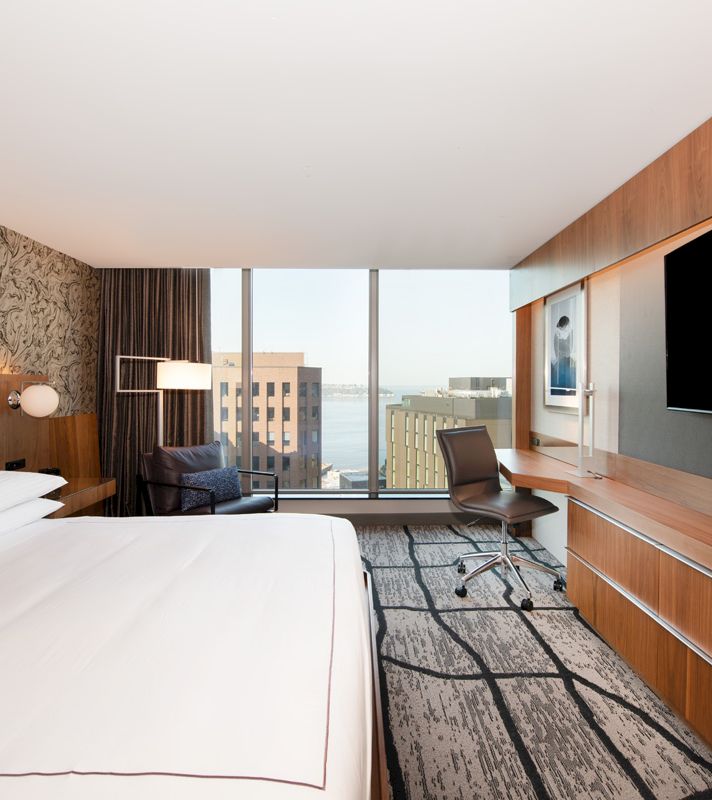 A hotel room with a bed, desk, chair, and TV in front of a window with a city view.