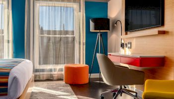 A modern room with a bed, desk, chair, TV, lamp, pouf, and window with sheer curtains. The decor features bright colors and sleek designs.