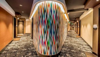 A brightly lit, modern hallway with patterned walls, doors leading to rooms, colorful abstract panel in the center, and carpeted flooring.