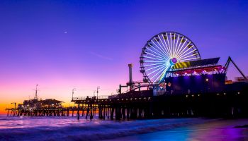 The image shows a vibrant pier at sunset with a lit Ferris wheel, colorful lights reflecting on the water, and a clear sky transitioning to night.