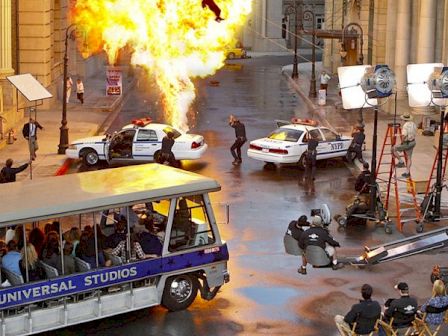 The image shows a film set with an explosion, a stunt person, police cars, a tram, and crew members working with cameras and lights.