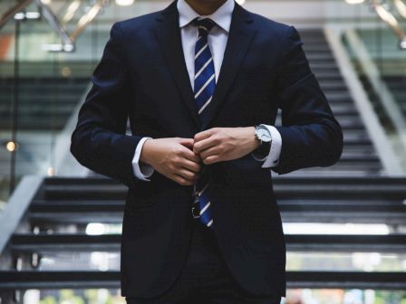 A person in a dark suit and striped tie is standing in front of stairs, adjusting their jacket with both hands, and wearing a watch.