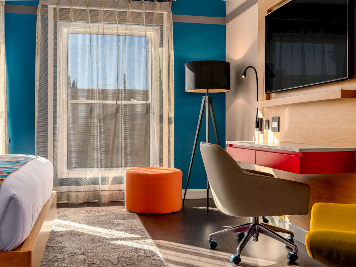 A modern hotel room with a bed, desk, chair, ottoman, TV, floor lamp, and large windows with sheer curtains, showcasing vibrant colors and sleek design.