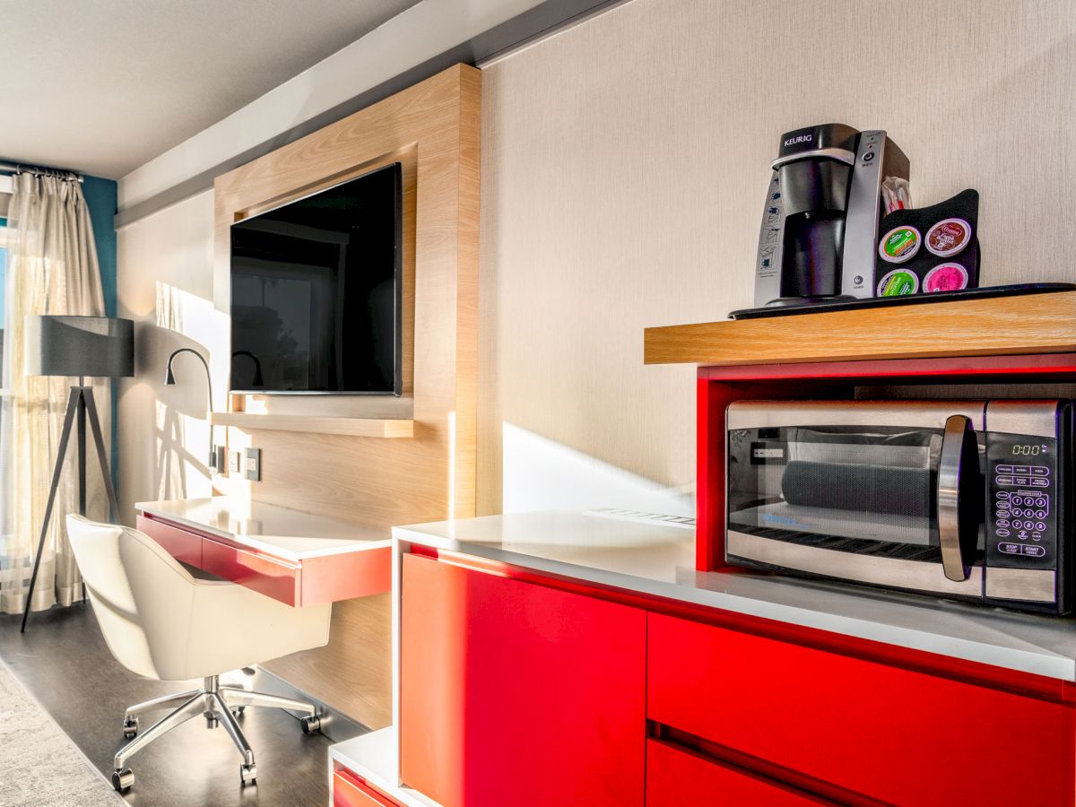 The image shows a modern hotel room with a desk, chair, wall-mounted TV, and kitchen area with a microwave and coffee maker.