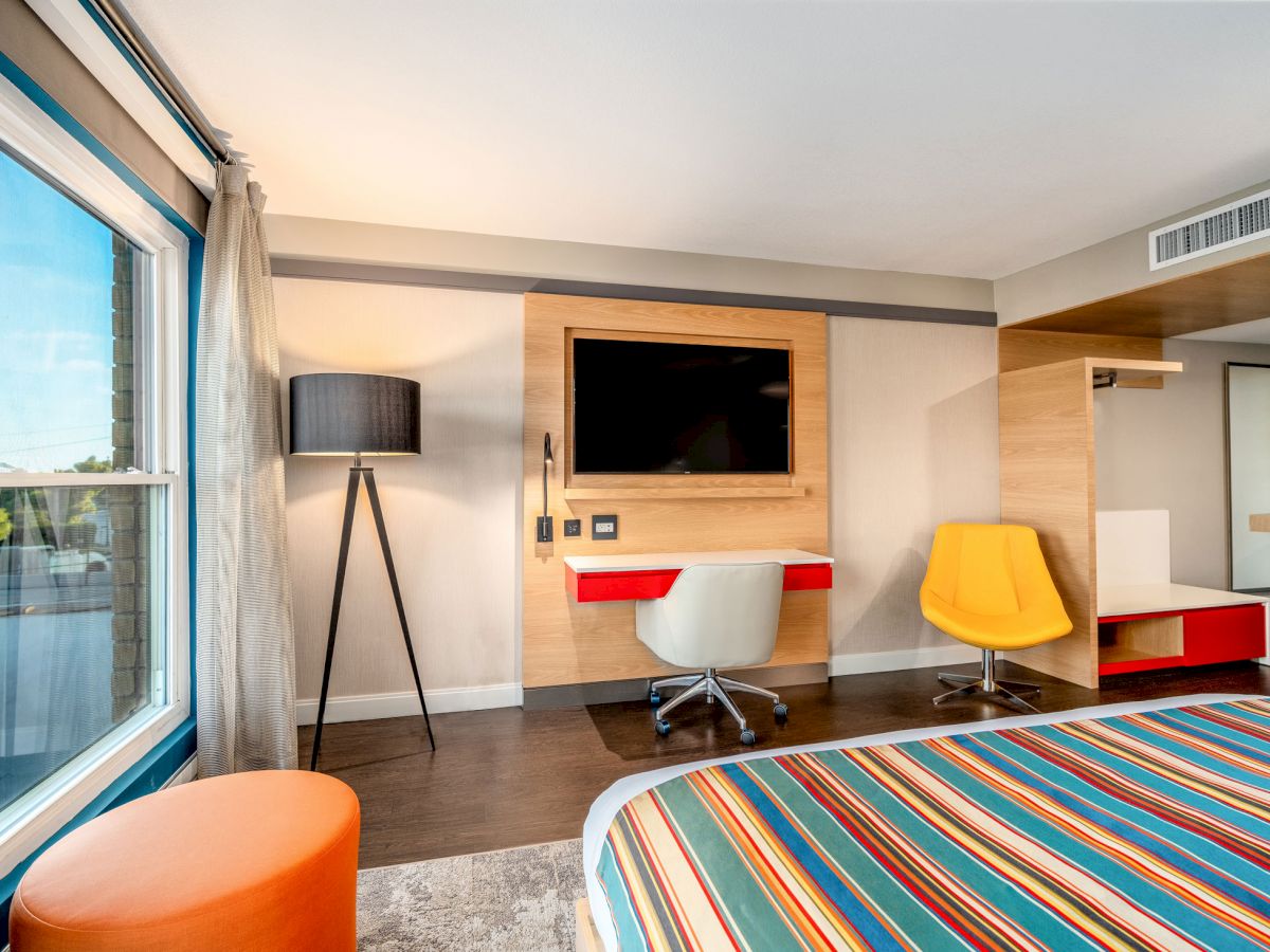 A modern hotel room with a large window, striped bed, TV, desk, white chair, yellow chair, black lamp, and orange ottoman.