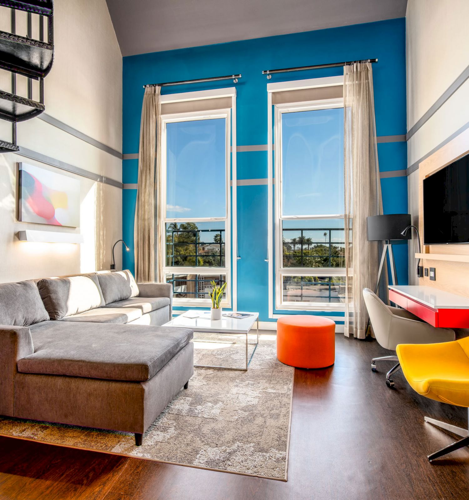 A modern living room with a gray sectional sofa, yellow chair, red desk, wall-mounted TV, and spiral staircase. There are tall windows with curtains.