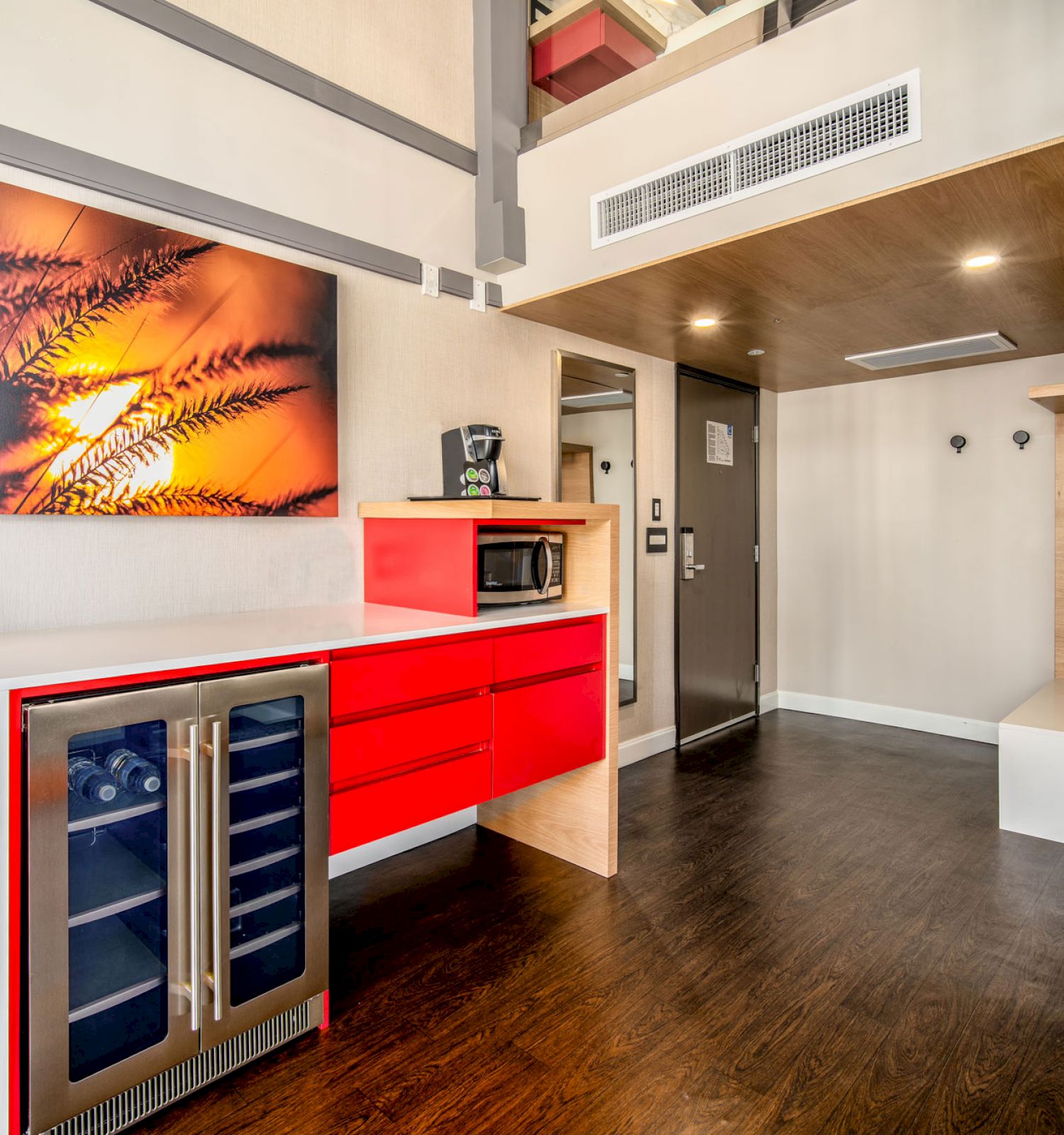 A modern hotel room with a kitchenette, including a wine cooler, microwave, and bright red cabinets, featuring wood flooring and wall art.