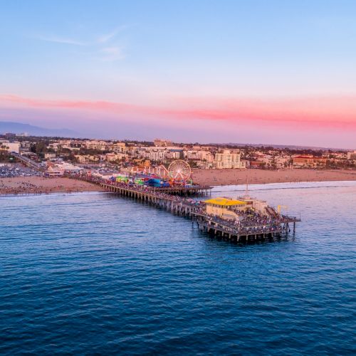 Aerial view of a bustling pier extending into the ocean, with a coastal city and sandy beach under a colorful sunset sky in the background.