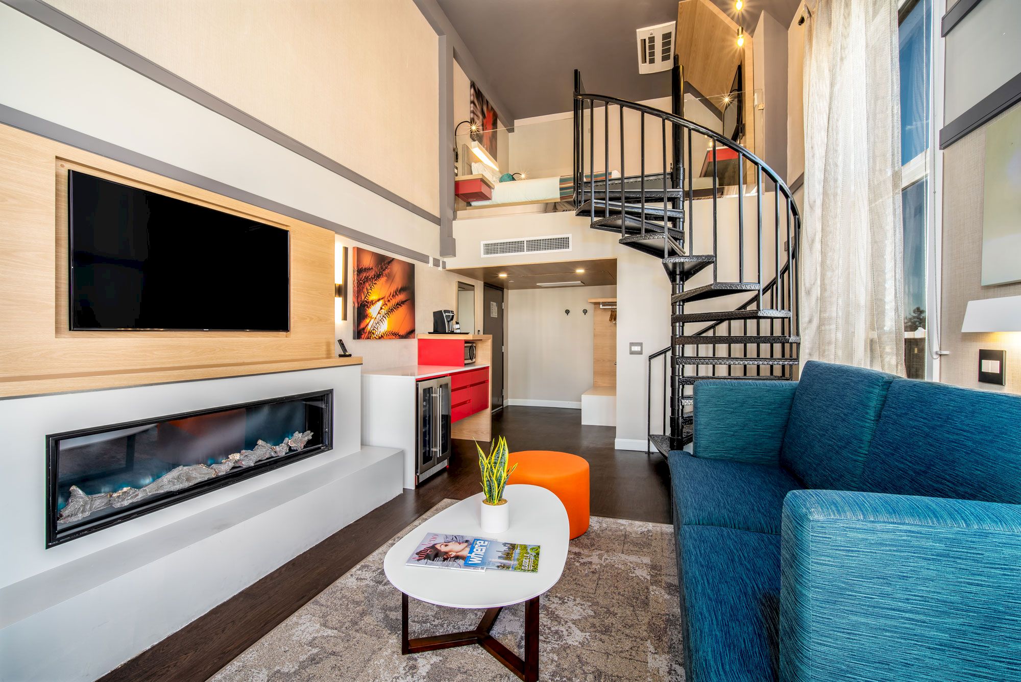 A modern living space features a blue sofa, a spiral staircase, a TV, a fireplace, a kitchenette with red accents, and a small coffee table.