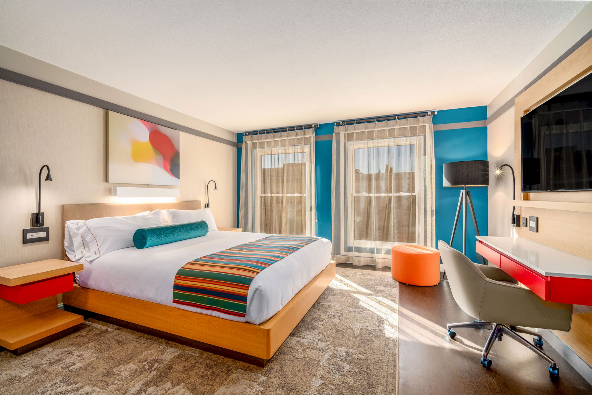 A modern hotel room with a large bed, colorful decor, desk, chair, TV, and natural light through large windows.